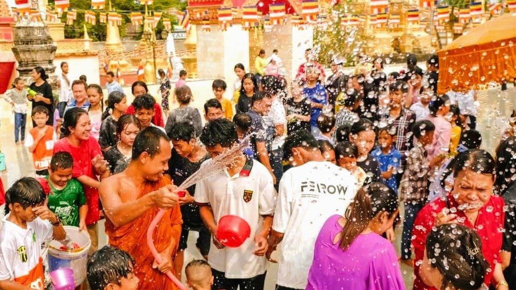 A monk in orange robes sprays a hose at a crowd of smiling people during a water fight at a Khmer pa